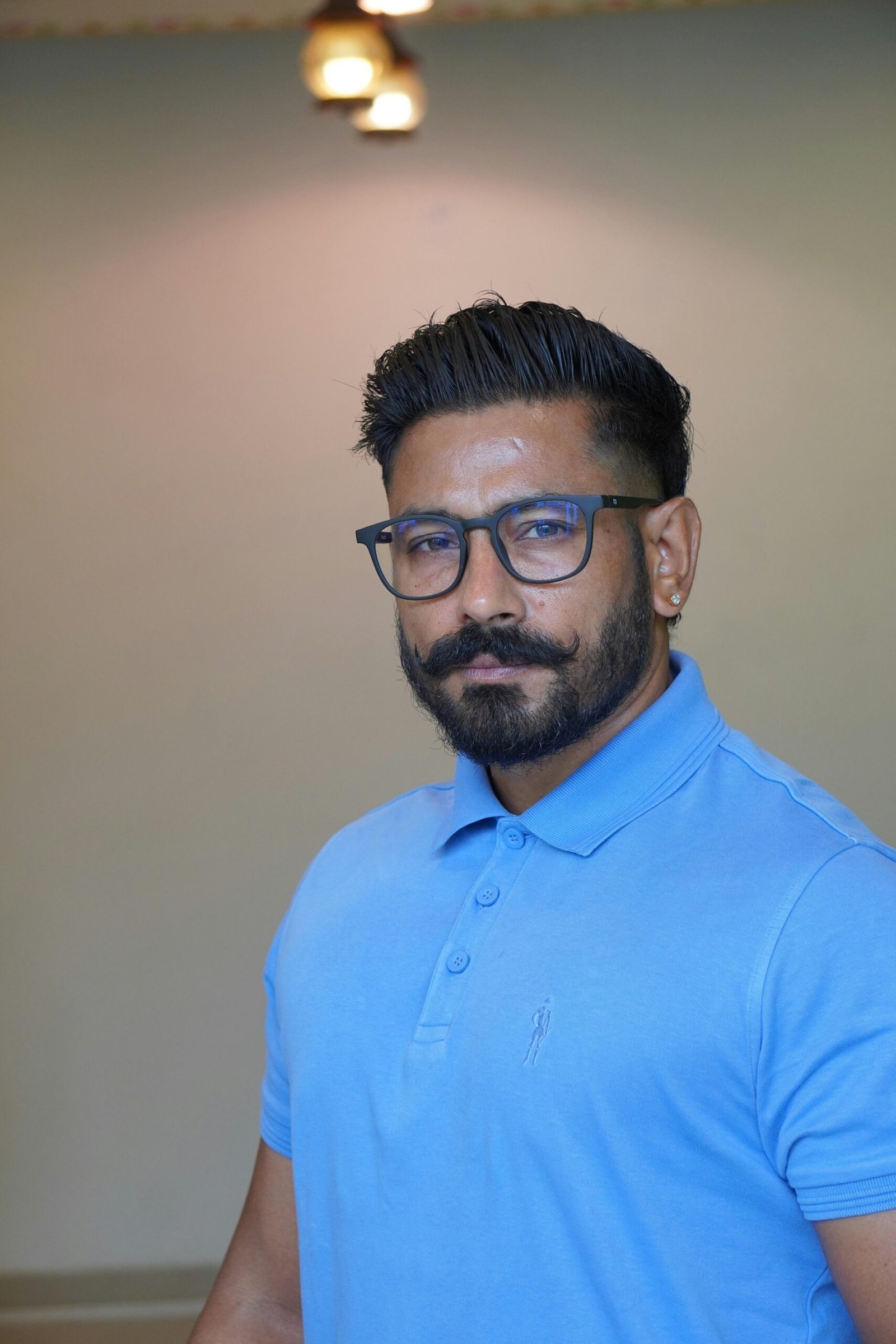 A confident man with eyeglasses and a mustache, wearing a blue polo shirt indoors.